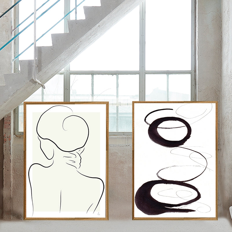 Two abstract art pieces in frames on a wall with a staircase and large window in the background.